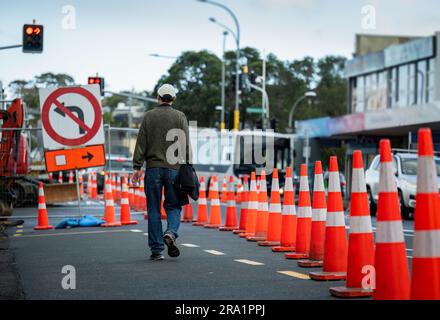 Homme marchant sur la route avec des cônes de circulation orange qui détournent le trafic. Bus et voitures sur la route en cours d'entretien. Auckland. Banque D'Images