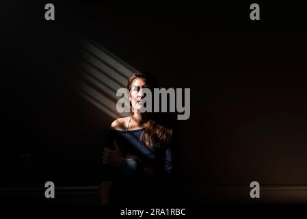 Portrait d'une femme assise dans une pièce sombre dans une bande lumineuse rayée. Banque D'Images