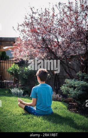 Vue arrière d'un garçon entre en position assise sur l'herbe dans un jardin méditant. Banque D'Images