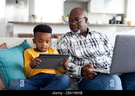 Heureux grand-père afro-américain et petit-fils assis sur le canapé, en utilisant tablette et ordinateur portable Banque D'Images