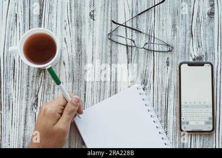 Homme écrit dans un bloc-notes avec sa main gauche dans un bloc-notes, maquette d'écran de téléphone. Verre et tasse de thé. Photo conceptuelle du jour de l'heureux gauche hander. Banque D'Images