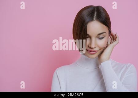 Jeune femme réfléchie, look timide, expression rêveuse, vêtements décontractés, pensées agréables. Isolé sur un arrière-plan rosé avec un espace vide pour la promotion. Banque D'Images