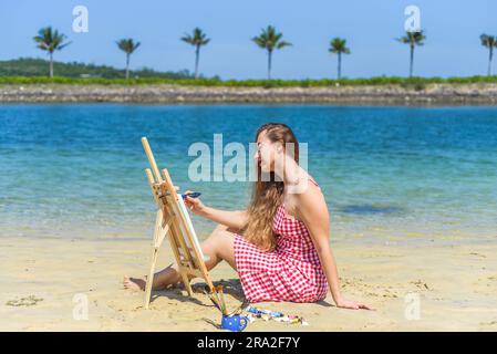 Jeune artiste femme assise à la plage avec un pot avec des pinceaux et un chevalet près de la mer et des palmiers en été Banque D'Images