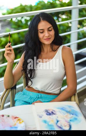 Femme brunette ukrainienne peignant le dessin à l'aquarelle d'une fille asiatique avec un pinceau sur une table avec de l'eau et des peintures Banque D'Images