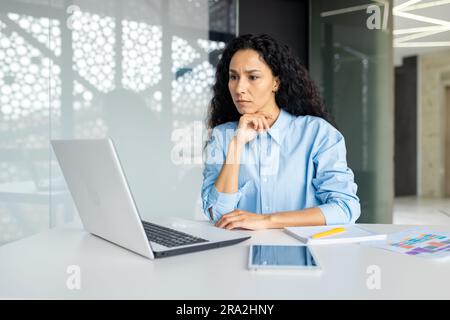Femme sérieuse de pensée travaillant au bureau sur le lieu de travail avec un ordinateur portable, femme hispanique confiante regardant la tâche technique, solution de brainstorming, femme d'affaires avec les cheveux bouclés regardant l'écran d'ordinateur portable Banque D'Images