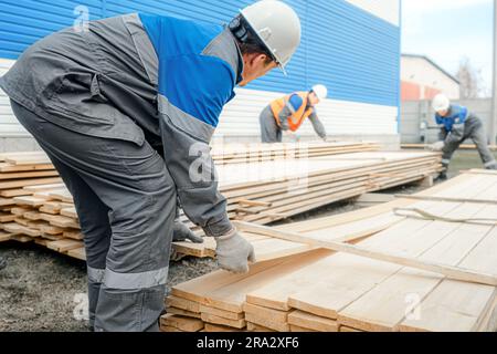 Les travailleurs en casques et combinaisons empilent des planches de bois à l'extérieur de la rue. Achat et vente de matériaux de construction. Arrière-plan industriel. Banque D'Images