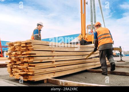 Deux travailleurs en casques et gilets de construction déchargent des planches en bois. Les déflecteurs empilent du bois à l'extérieur en plein air. Travaux de chargement et de déchargement. Flux de travail authentique. Banque D'Images