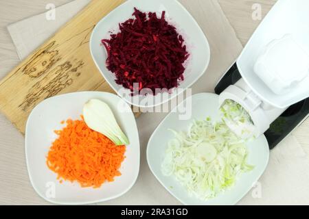 Carottes, betteraves et oignons dans un coupe-légumes sur une table de cuisine. Les légumes hachés tombent dans des bols. Aliments sains faits maison. Banque D'Images