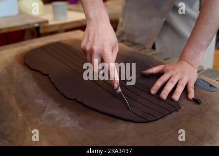 femme coupe la céramique dans l'atelier de poterie travaillant vase en céramique brute et argile pour faire pot. Atelier de poterie à la lumière naturelle raclage, lissage Banque D'Images