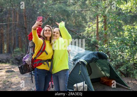 Maman avec sa fille lors d'un appel vidéo en ligne dans un camp forestier. Ils se tiennent près de la tente et communiquent avec leur père par liaison vidéo. Maternité et tr Banque D'Images