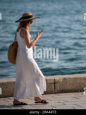 élégante jeune femme en longue robe d'été blanche et chapeau de soleil de paille marchant le long du front de mer en utilisant smartphone le soir d'été dans la méditerranée. Banque D'Images