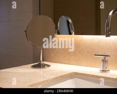 Petit miroir de maquillage rond pliant près du lavabo avec dessus en marbre et robinet en acier près de la grande fenêtre murale en verre avec éclairage caché. Mod Banque D'Images