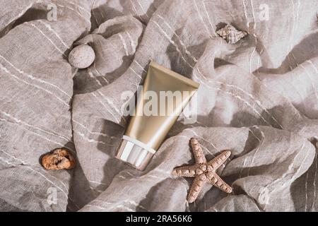 Tube crème dorée et coquillages sur tissu de coton beige au soleil. Vue de dessus, plan d'agencement, maquette. Banque D'Images