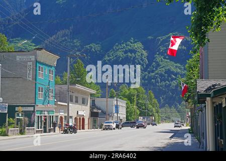 Vieilles maisons en bois de l'époque pionnière dans la rue principale du village de Stewart, drapeau canadien et paysages de montagne verdoyants, Colombie-Britannique Banque D'Images