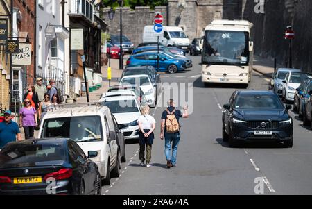 Des gens, probablement des touristes, marchant le long d'une route très fréquentée malgré qu'il y ait des voitures, dans une ville britannique en Angleterre, au Royaume-Uni. Banque D'Images