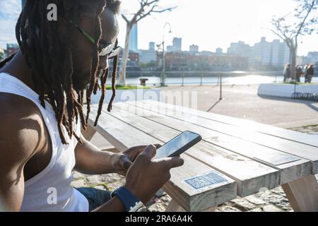 Homme africain avec des dreadlocks, des lunettes de soleil, assis à l'extérieur d'un restaurant à Puerto Madero Argentine au coucher du soleil scannant le code QR avec son téléphone pour voir Banque D'Images