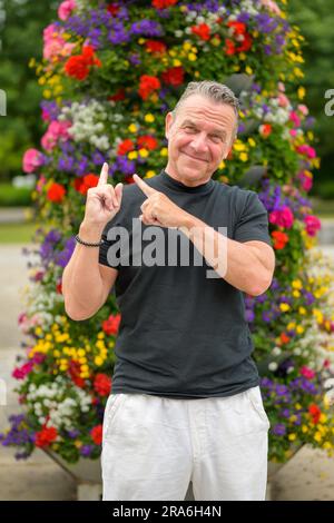 Portrait de trois quarts de longueur d'un vieil homme attrayant pointant ses doigts vers l'arrière sur une grande tour de fleurs Banque D'Images