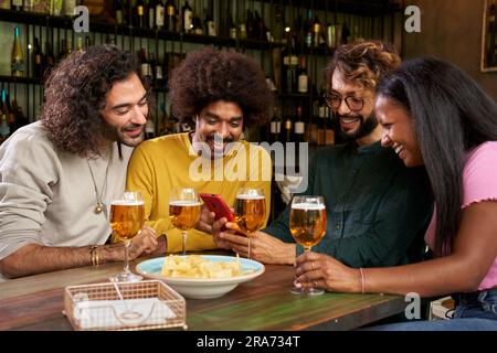 Groupe de jeunes regardant au mobile dans un bar. Ces quatre amis multiethniques sourient Banque D'Images