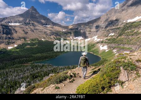 Homme caucasien mûr marchant sur un sentier vers une vallée avec un lac entouré de montagnes accidentées, parc national de Glacier, Montana Banque D'Images