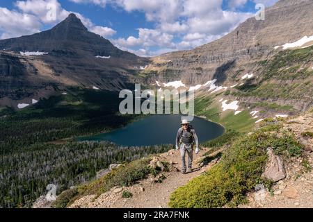 Homme caucasien mûr marchant sur un sentier sortant d'une vallée avec un lac entouré de montagnes accidentées, parc national de Glacier, Montana Banque D'Images