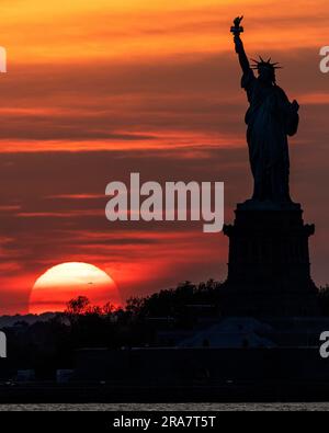 Silhouette de la Statue de la liberté avec un immense soleil couchant en arrière-plan et un ciel et des nuages orange rouge et jaune Banque D'Images