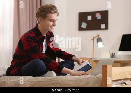 Formation en ligne. Un adolescent souriant lisant le livre près d'un ordinateur portable sur le lit à la maison Banque D'Images