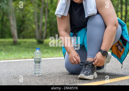 Une jeune femme en surpoids s'arrête pour attacher sa chaussure pendant sa course d'exercice matinal sur une piste de course d'un parc local Banque D'Images