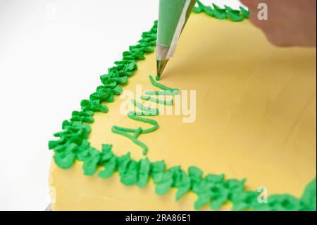 Femme écrivant avec un sac de pâtisserie sur un gâteau d'anniversaire vert et jaune isolé sur fond blanc. Banque D'Images