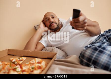 Portrait d'un adulte ennuyé regardant la télévision à la maison et allongé sur le lit avec une pizza, espace de copie Banque D'Images