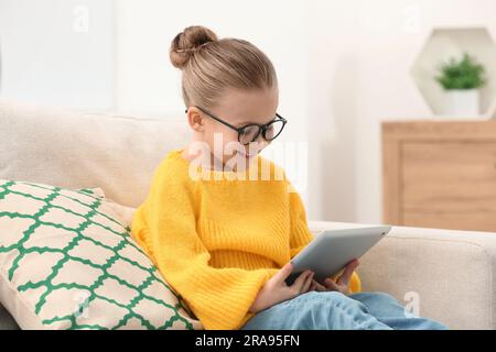 Jolie petite fille dans des verres avec une tablette sur le canapé à l'intérieur Banque D'Images