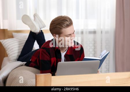 Formation en ligne. Un adolescent souriant lisant le livre près d'un ordinateur portable sur le lit à la maison Banque D'Images