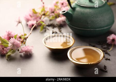 Cérémonie traditionnelle. Tasses de thé filtre, théière et branche de l'arbre de sakura sur table grise, en gros plan Banque D'Images