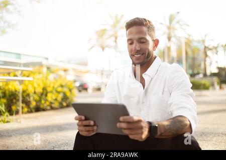 Homme d'affaires joyeux et joyeux lisant des informations sur une tablette numérique, assis à l'extérieur près du bureau pendant la pause, espace libre Banque D'Images