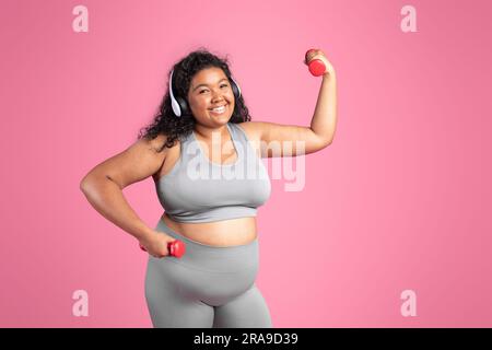 Bonne femme afro-américaine en vêtements de sport, casque sans fil faisant de l'exercice avec des haltères, espace libre Banque D'Images