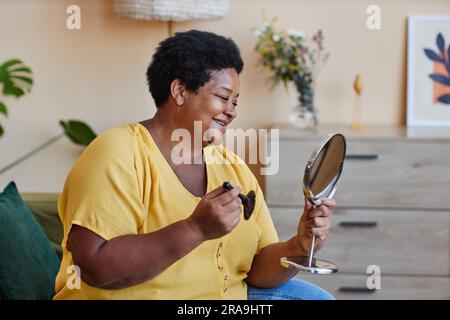 Une femme noire d'âge heureux se regardant dans le miroir avec le sourire tout en appliquant le maquillage avec la brosse de visualisation devant l'appareil photo dans le salon Banque D'Images