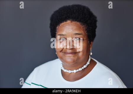 Portrait d'une femme afro-américaine âgée en chandail blanc, collier de perles et boucles d'oreilles regardant la caméra avec le sourire contre le mur gris en studio Banque D'Images