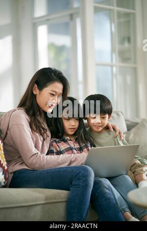 une jeune mère asiatique et deux enfants assis sur un canapé de famille à la maison en utilisant un ordinateur portable ensemble Banque D'Images