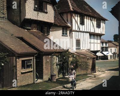 Une petite fille avec un arc dans ses cheveux roule son vélo dans un coin de la vieille ville pittoresque de Thaxted, Essex, Angleterre, avec ses vieilles maisons de chaume à côté de la salle de guilde Date: 1950s Banque D'Images