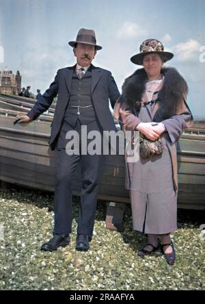 Un couple d'âge moyen debout devant quelques bateaux sur une plage de galets. Date: C.1920 Banque D'Images