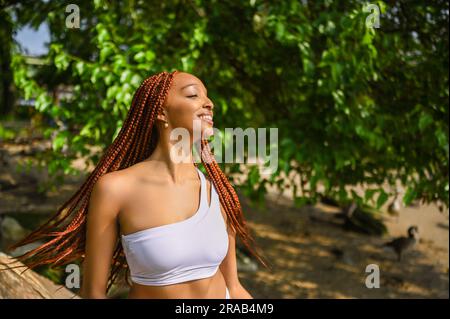 Portrait extérieur de la belle nature jeune afro-américaine femme long rouge braid cheveux style et dents blanches parfaites sourire, posant en maillot de bain à la journée ensoleillée d'été avec le feuillage vert plage arrière-plan Banque D'Images