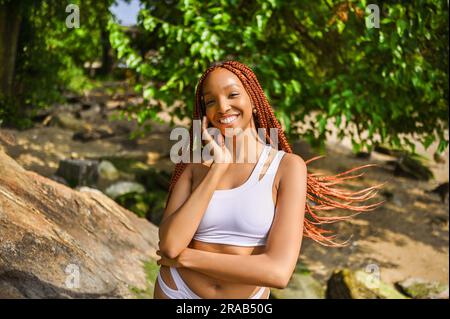 Portrait extérieur de la belle nature jeune afro-américaine femme long rouge braid cheveux style et dents blanches parfaites sourire, posant en maillot de bain à la journée ensoleillée d'été avec le feuillage vert plage arrière-plan Banque D'Images