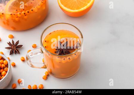 Thé d'automne à l'argousier avec de l'orange dans une tasse de verre et théière avec baies fraîches, cannelle et romarin sur table blanche Banque D'Images