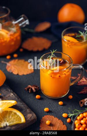 Thé chaud d'automne avec baies d'argousier, orange, cannelle et romarin dans des tasses en verre et théière sur fond sombre Banque D'Images