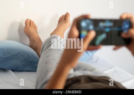 Adolescent pieds nus avec jambes croisées reposant sur un oreiller à l'aide d'un téléphone allongé sur le lit. Un adolescent jouant à des jeux vidéo dans sa chambre en milieu de matinée. Banque D'Images