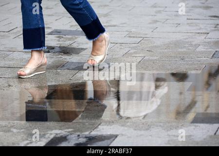 Jambes de femme en jeans et chaussures sur trottoir mouillé, femme courir dans une rue avec flaque après la pluie Banque D'Images