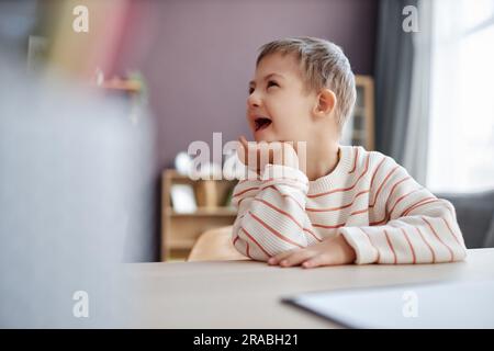 Vue latérale portrait d'un petit garçon heureux avec le syndrome de Down assis au bureau à l'école et rire, espace de copie Banque D'Images