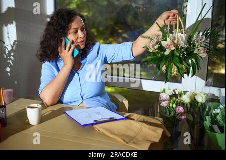Femme confiante fleuriste entrepreneur parlant sur téléphone mobile, tenant un panier en osier avec arrangement de fleurs, discutant des commandes pour les fêtes spéciales l Banque D'Images