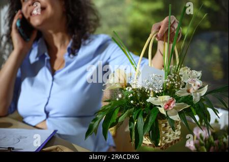 Accent sélectif sur un beau arrangement de fleurs avec des orchidées et des plantes fraîches dans le panier en osier sur la femelle floue fleuriste discutant de la livraison le o Banque D'Images