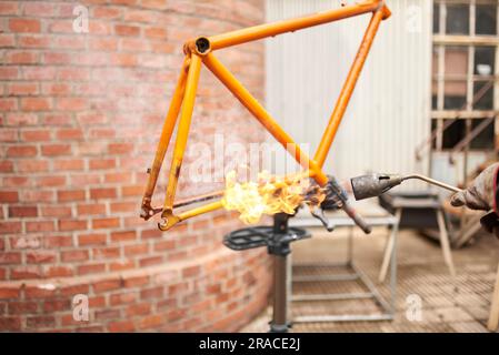 Personne méconnaissable utilisant un chalumeau pour enlever la peinture d'un cadre de vélo orange qui est en cours de rénovation dans son atelier. Banque D'Images