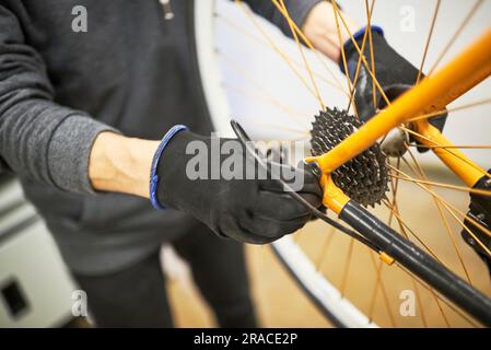 Entretien d'une bicyclette : mains d'une personne méconnaissable utilisant des gants pour démonter la roue arrière d'une moto orange dans son atelier de réparation. Banque D'Images